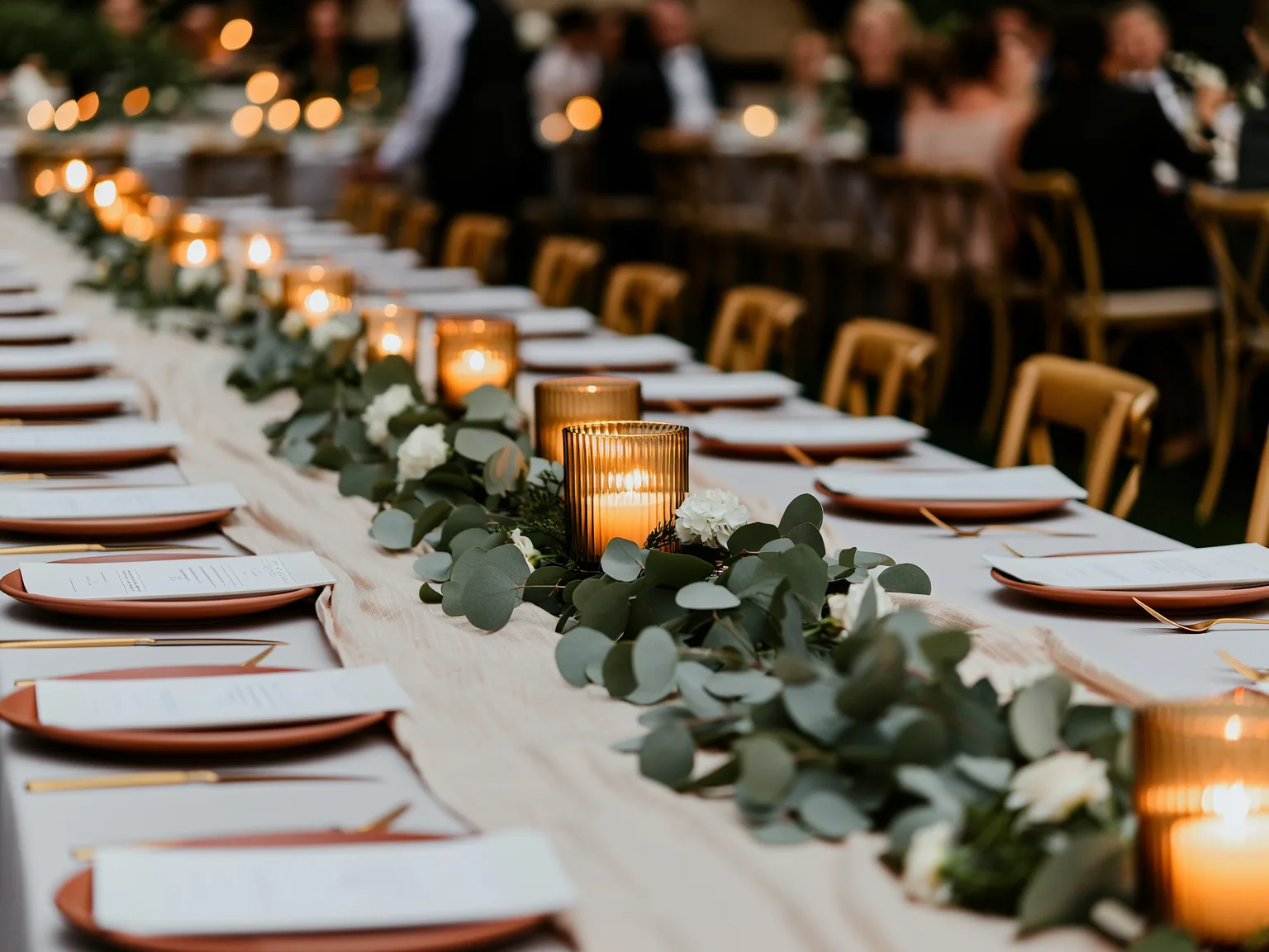 Mesa larga de boda al atardecer, con velas encendidas, camino de eucalipto, platos y menús puestos